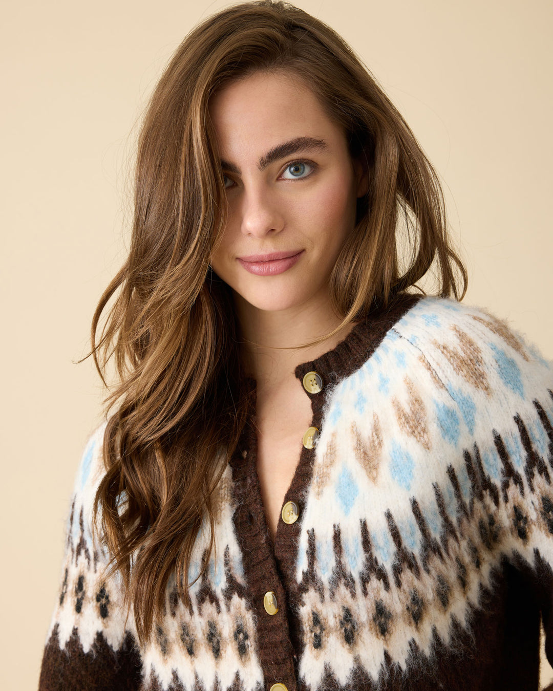 A woman with long wavy brown hair wearing a brown, cream, and light blue patterned knit cardigan with gold buttons, set against a solid beige background.