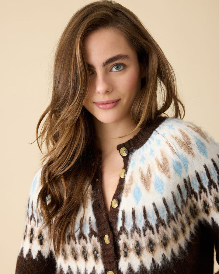 A woman with long wavy brown hair wearing a brown, cream, and light blue patterned knit cardigan with gold buttons, set against a solid beige background.